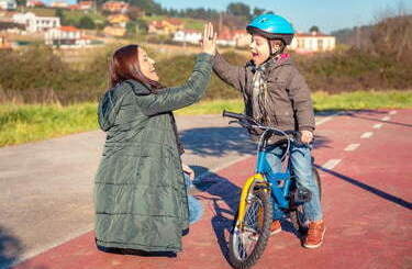 Mam z synem przyb ija piątkę, Syn siedzi na rowerze i ma założony kask.